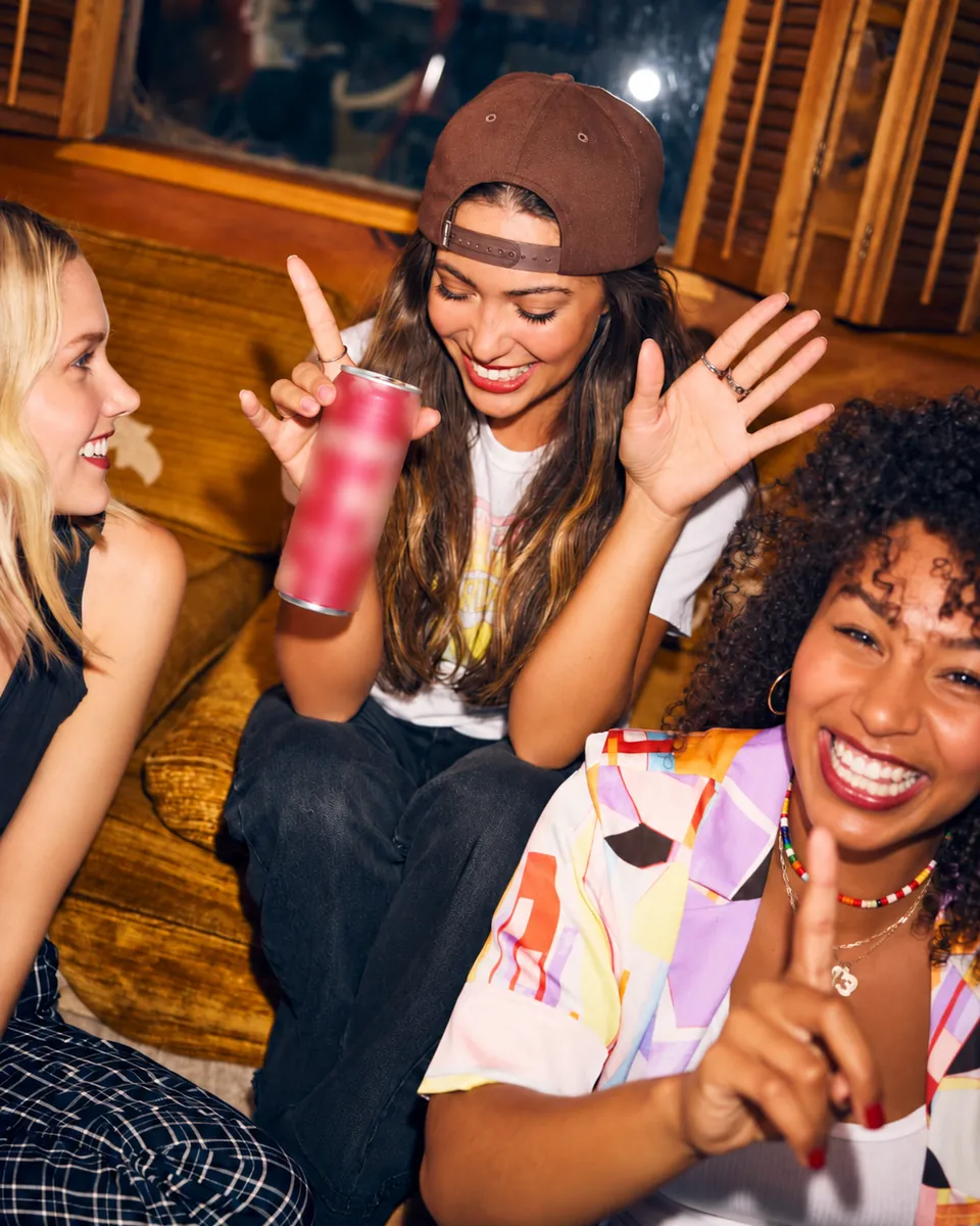 Três amigas rindo juntas em um sofá, uma delas segurando uma lata.